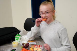 Білява дівчина в круглих окулярах у сірій сорочці їсть шматок пепероні