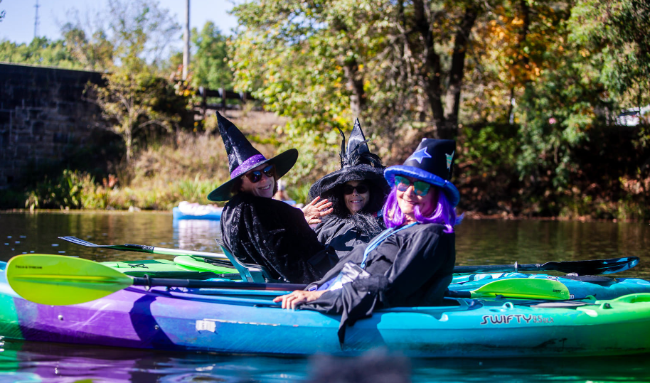 3 white women sitting in kayaks on a river dressed up as witches
