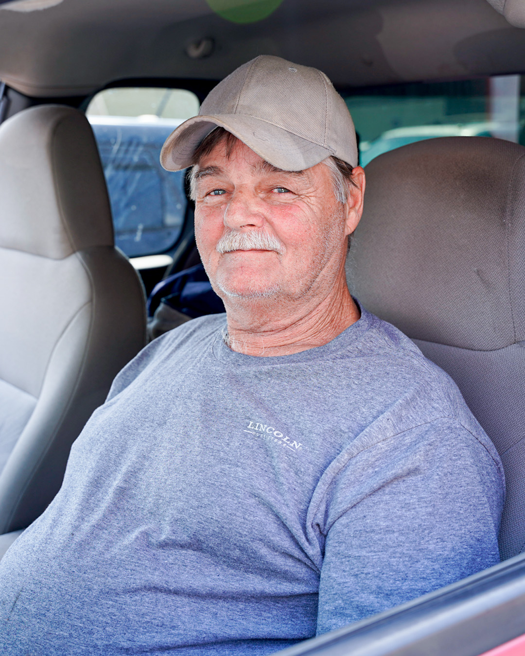 Old white man sitting in his car wearing a tan baseball cap and a grey t-shirt