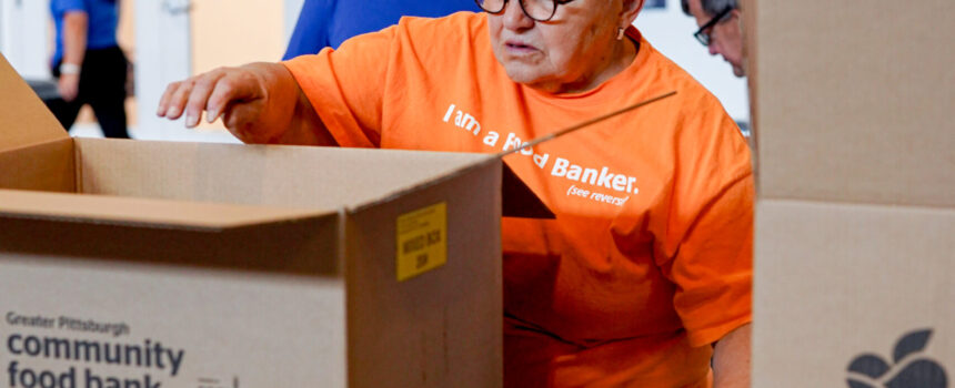 older white woman with white hair wearing an orange shirt, reaching in a brown box