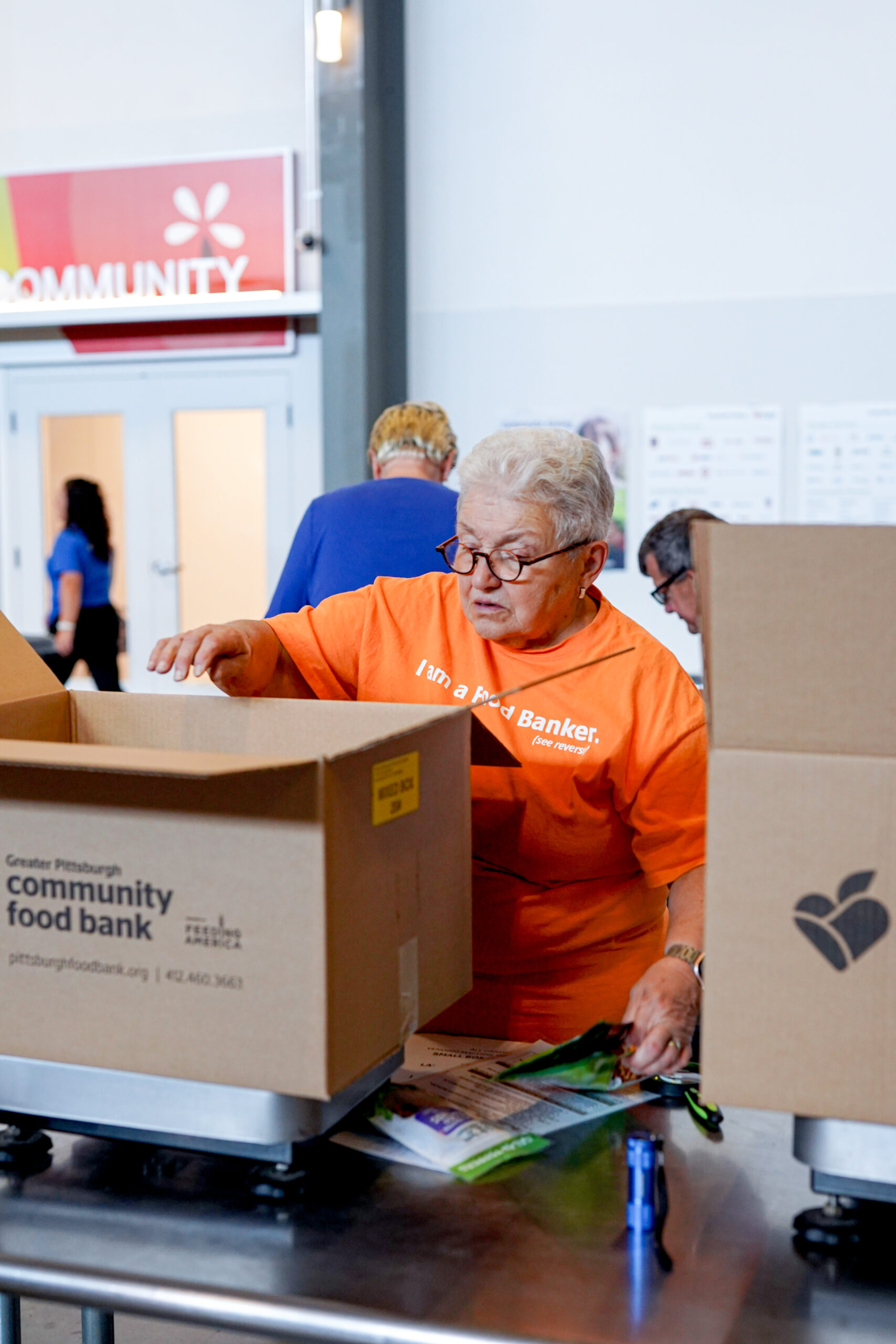 older white woman with white hair wearing an orange shirt, reaching in a brown box