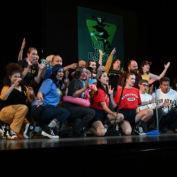 Group of people wearing colorful shirts in the spotlight on a black stage.